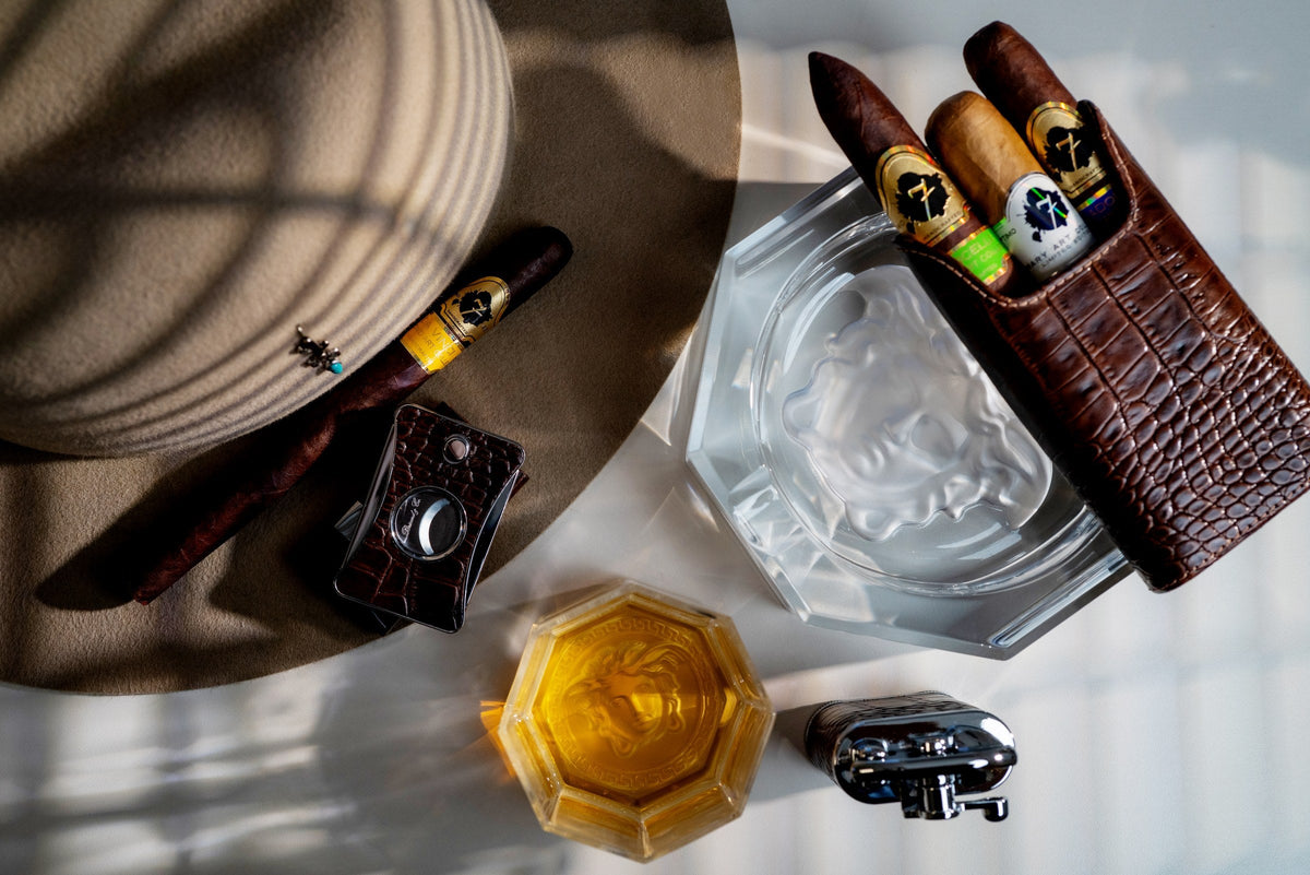 Premium cigar resting on a felt hat with a leather cigar case, crystal ashtray, cutter and whiskey